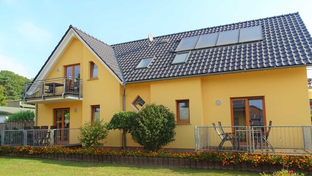 Ein zweistöckiges gelbes Haus mit einem Ziegeldach, einem Balkon und einem kleinen Garten mit Blumen.