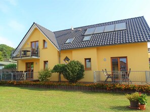 Une maison jaune à deux étages, avec un toit en tuiles, un balcon et un petit jardin fleuri.