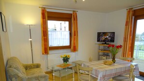 A cozy living room with a sofa, a glass coffee table, a dining table with chairs, a TV, and a window with curtains. - Vacation apartment 2 - Kos - Holiday home Ohm (Koserow)