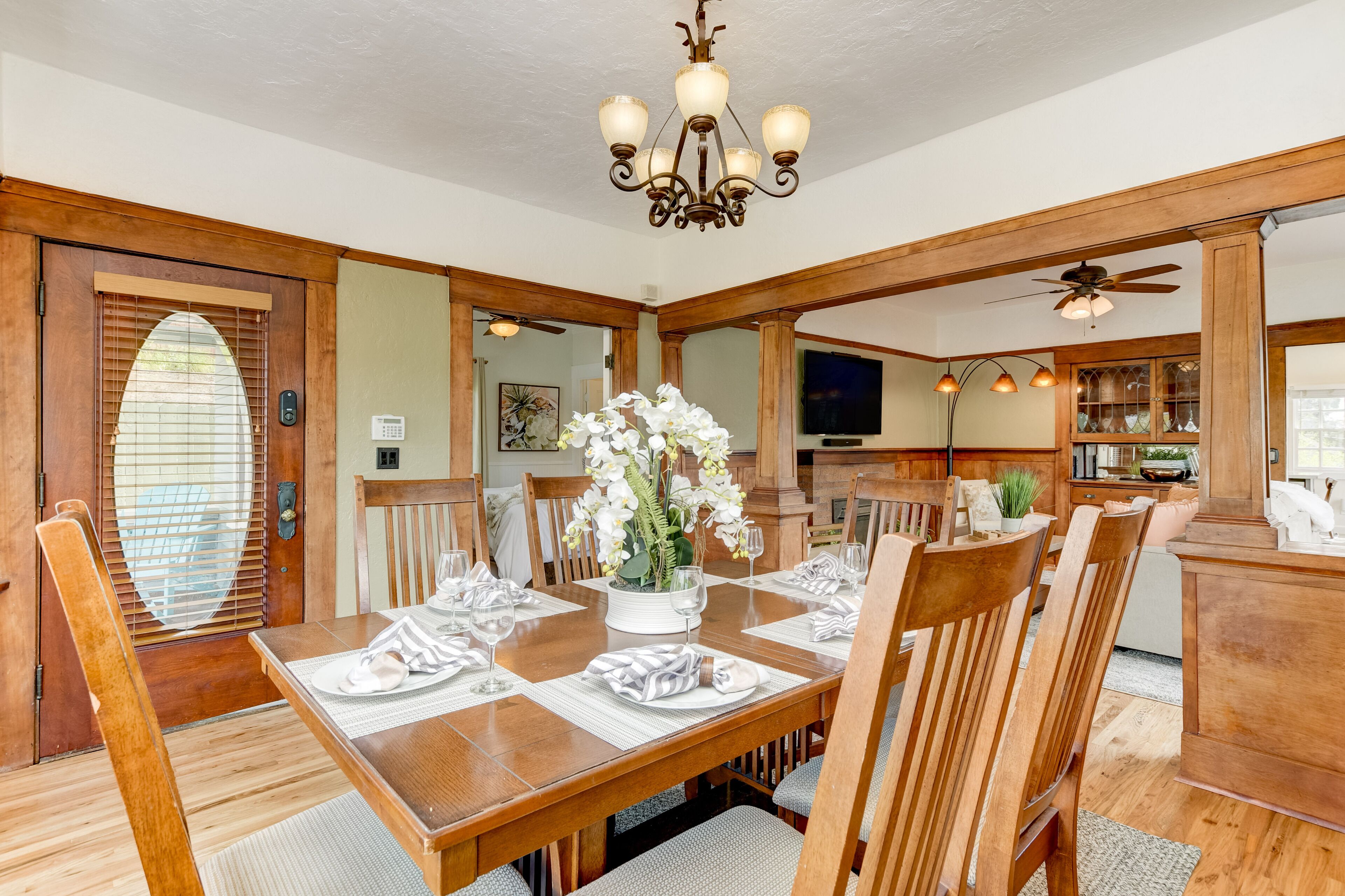 A dining area with a wooden table set for six, surrounded by chairs. There is a chandelier above the table and a ceiling fan in the background.