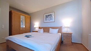 A neatly made bed with white linens, two bedside lamps, a wooden headboard, a framed artwork on the wall, and a wooden wardrobe. - An den Boddenwiesen III (Wustrow)