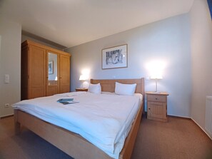 Ein ordentlich bezogenes Bett mit weißen Leinen, zwei Nachttischlampen, ein hölzernes Kopfende, ein gerahmtes Kunstwerk an der Wand und ein Holzschrank.