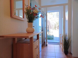 Ein Flur mit einem Holzkonsolentisch, einer Blumenvase, einem Spiegel und einer zur Außenseite führenden Tür.