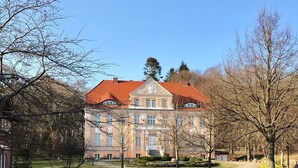 Un edificio grande e histórico con tejado de tejas rojas y arquitectura clásica, rodeado de un césped cuidado y árboles.