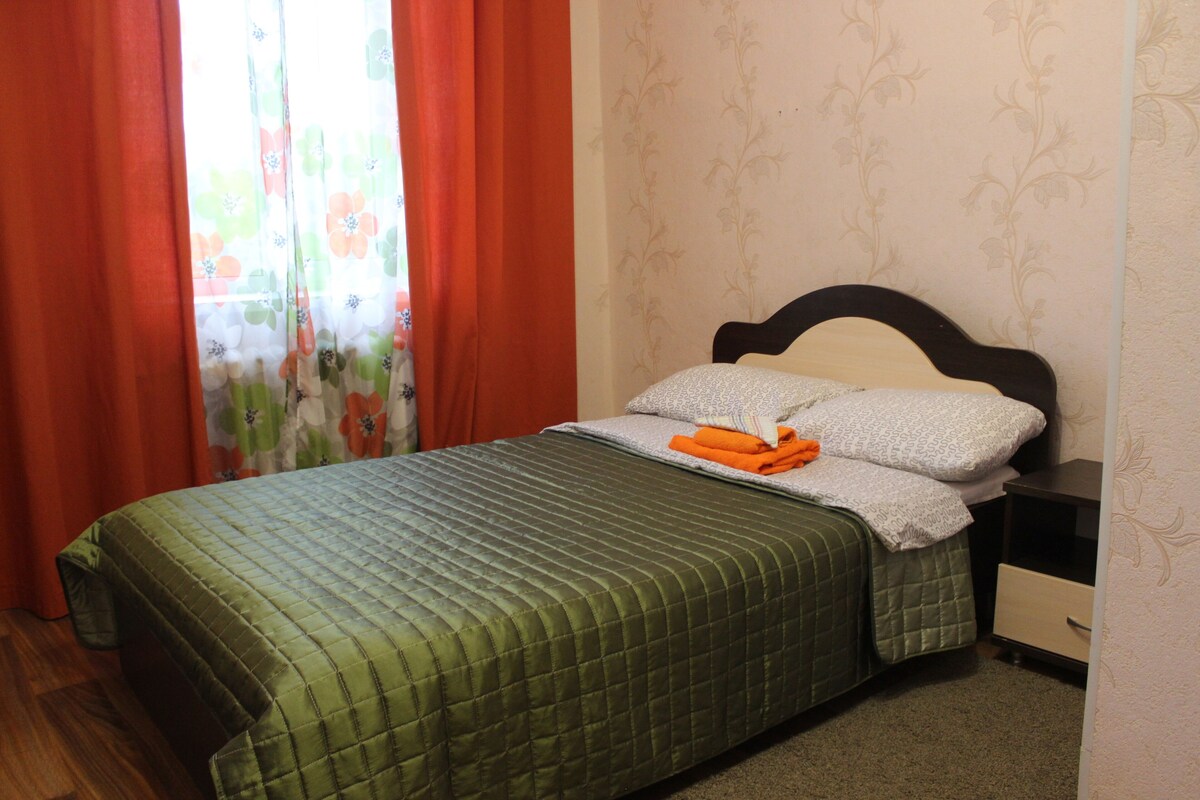 Una cama bien hecha con colcha verde, dos almohadas y toallas naranjas dobladas.