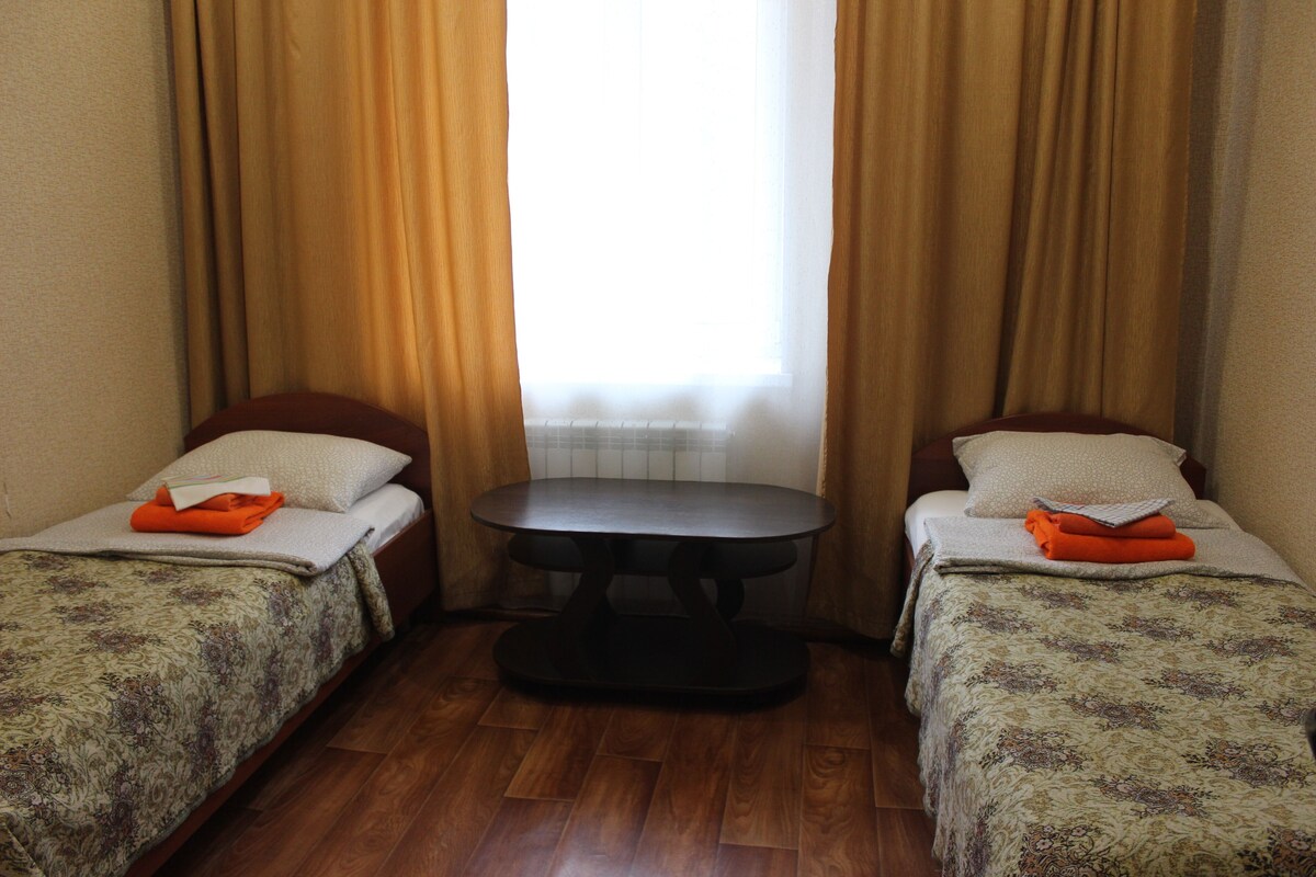 Una habitación con dos camas, una mesita y un ventanal con cortinas.