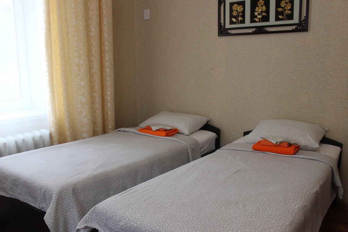 Dos camas individuales con ropa de cama gris y toallas naranjas, una ventana con cortina, y un cuadro con flores colgado en la pared.