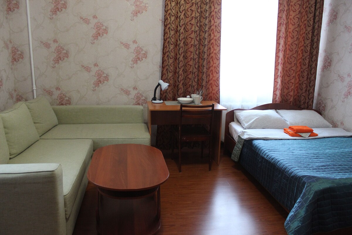 Una habitación con un sofá verde, una mesa de madera con una lámpara, una cama con una colcha azul y una ventana con cortinas estampadas.
