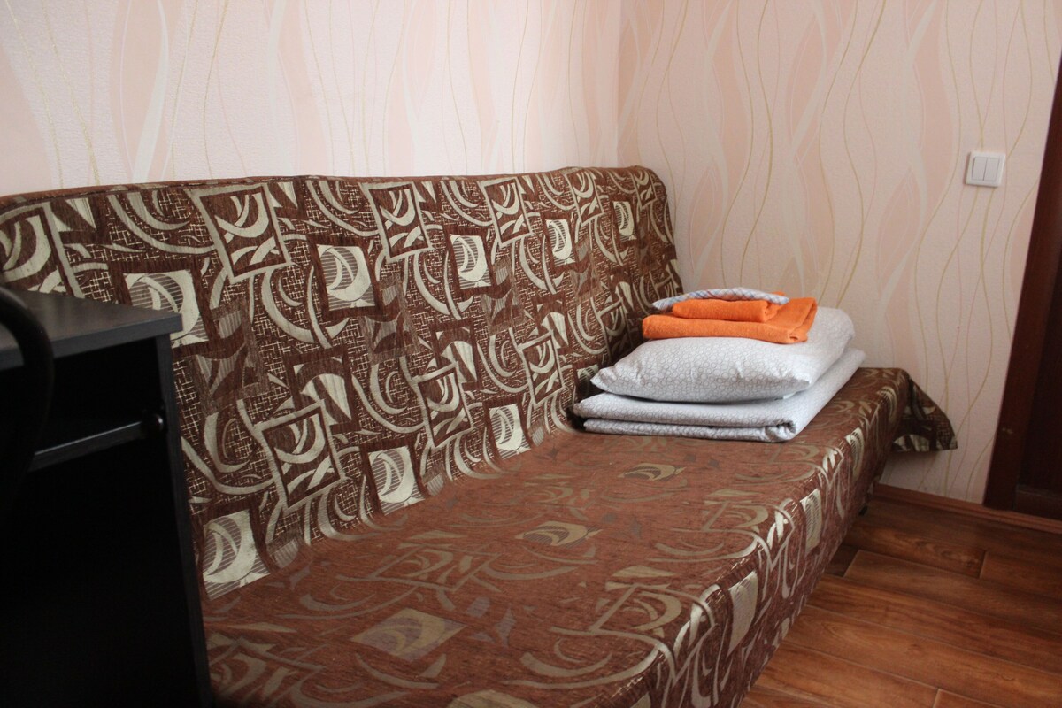 Un sofá con estampado, mantas dobladas y almohadas, un mueble oscuro a la izquierda y un suelo de madera.