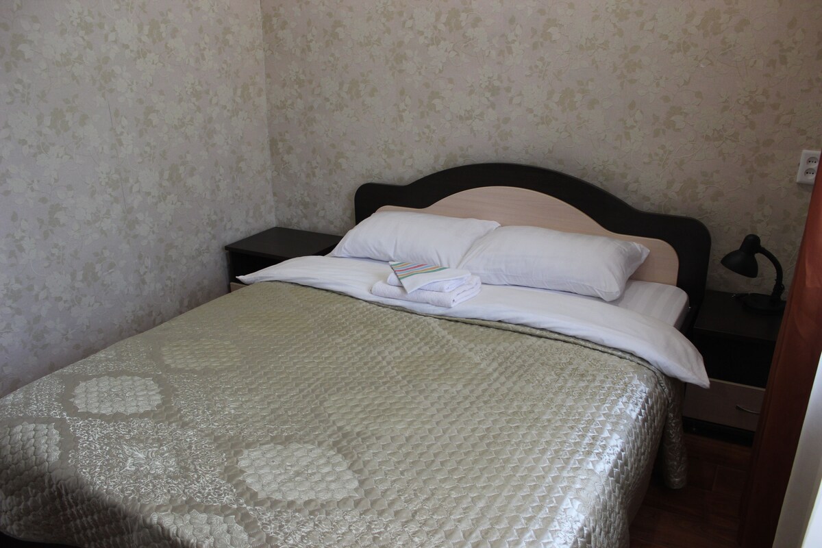 Una cama bien hecha con una colcha estampada, una almohada y una toalla doblada sobre la mesita de noche.