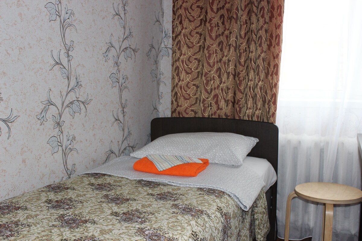 Una cama bien tendida con una colcha de motivos florales, una sola almohada naranja y un taburete de madera a un lado.