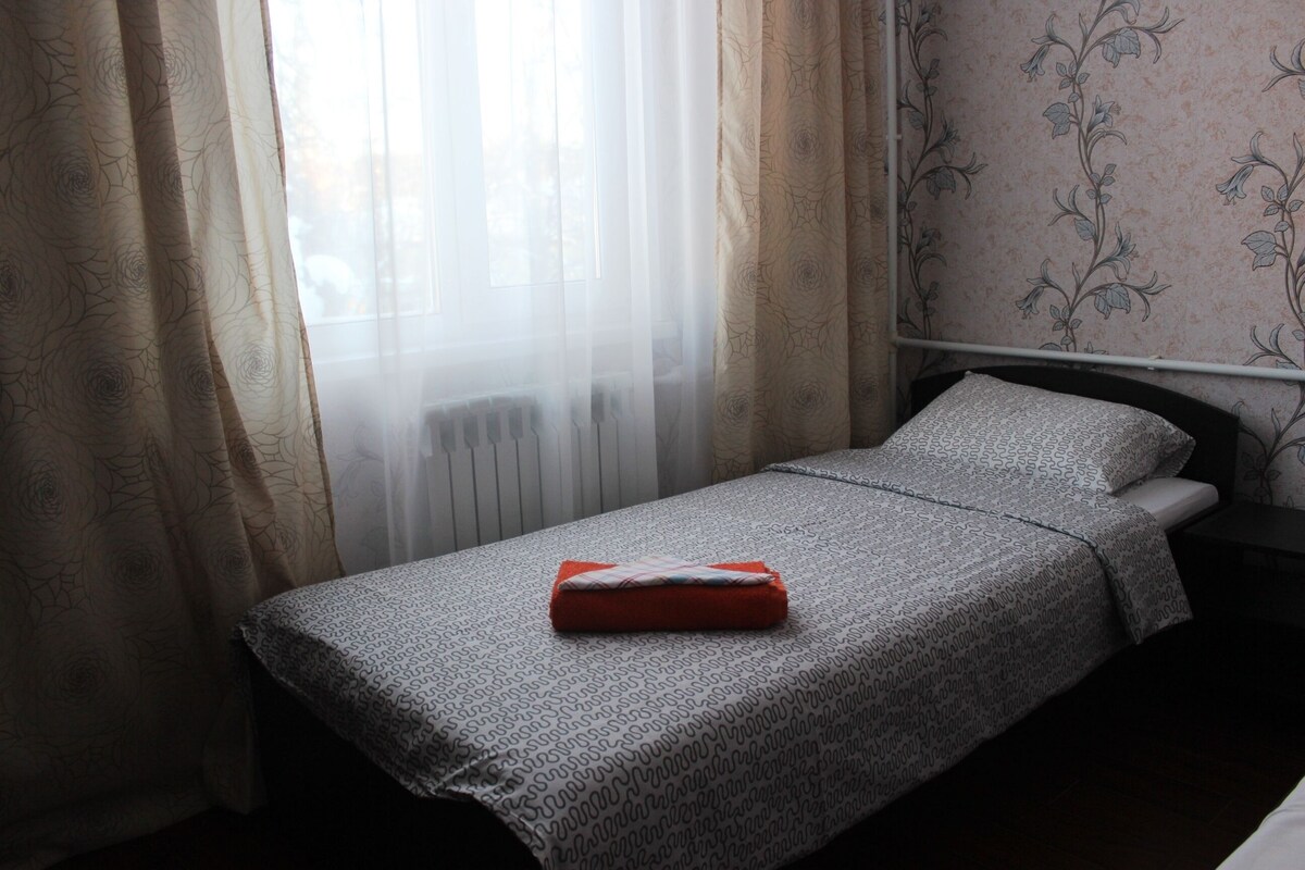 Una cama individual con una colcha estampada, un almohadón rojo y cortinas de gasa.