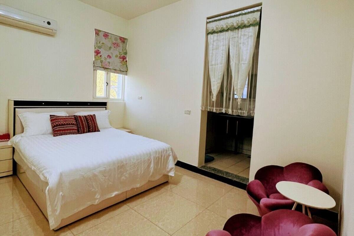 A bedroom with a bed, a nightstand, a window with curtains, a mirror, and two red chairs.