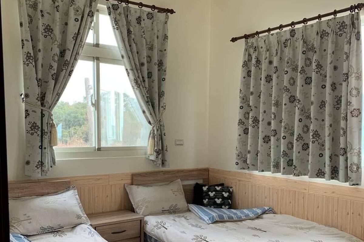 A room with two beds, a window with curtains, a bedside table, and a nightstand.