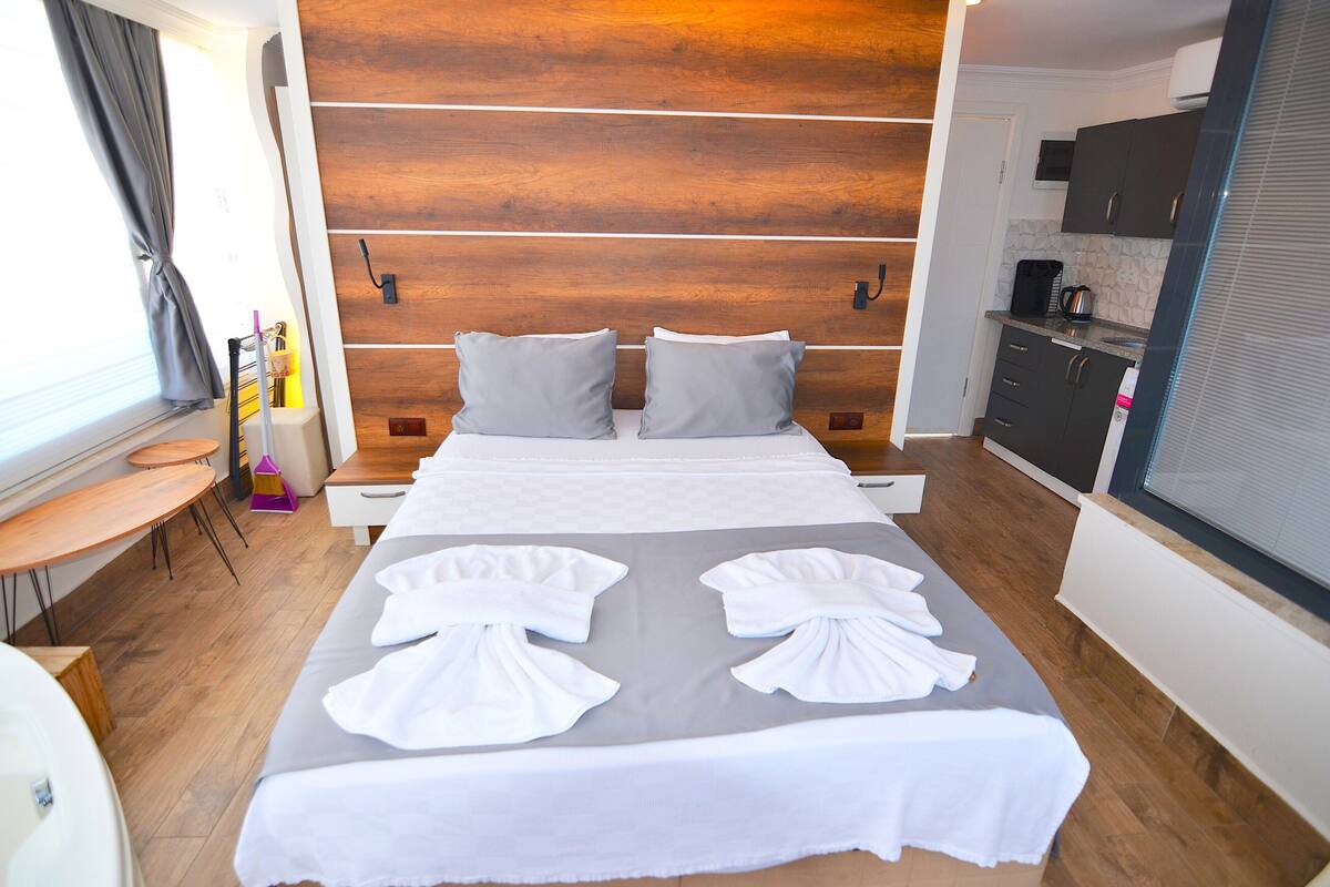 Una habitación de hotel con cabecera de madera, cama con ropa y toallas blancas, una mesita con una silla y una zona de kitchenette.