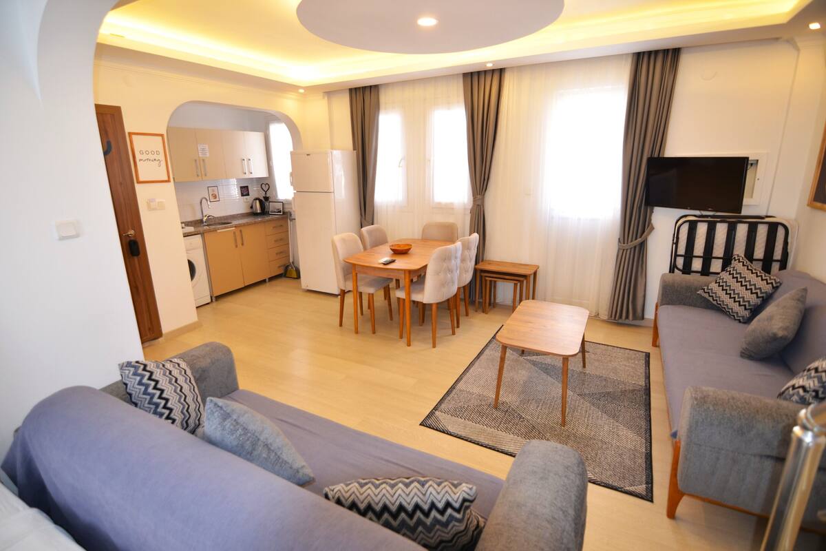 Una sala moderna con un sofá, una mesa de comedor con sillas, un área de cocina y un televisor montado en la pared.