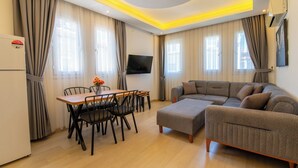 A modern living room with a sofa, a dining table with chairs, a flat-screen TV, and a ceiling light fixture. - Mykonut Apart Hotel (Fethiye)