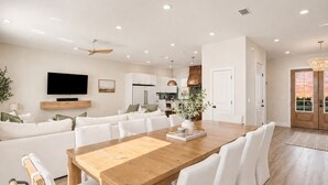 Amplia sala de estar con una gran mesa de comedor de madera, sofás blancos y un televisor de pantalla plana montado en la pared.