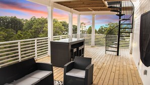 Una terraza techada al aire libre con mobiliario de mimbre, una escalera en espiral y vistas a árboles y al cielo.