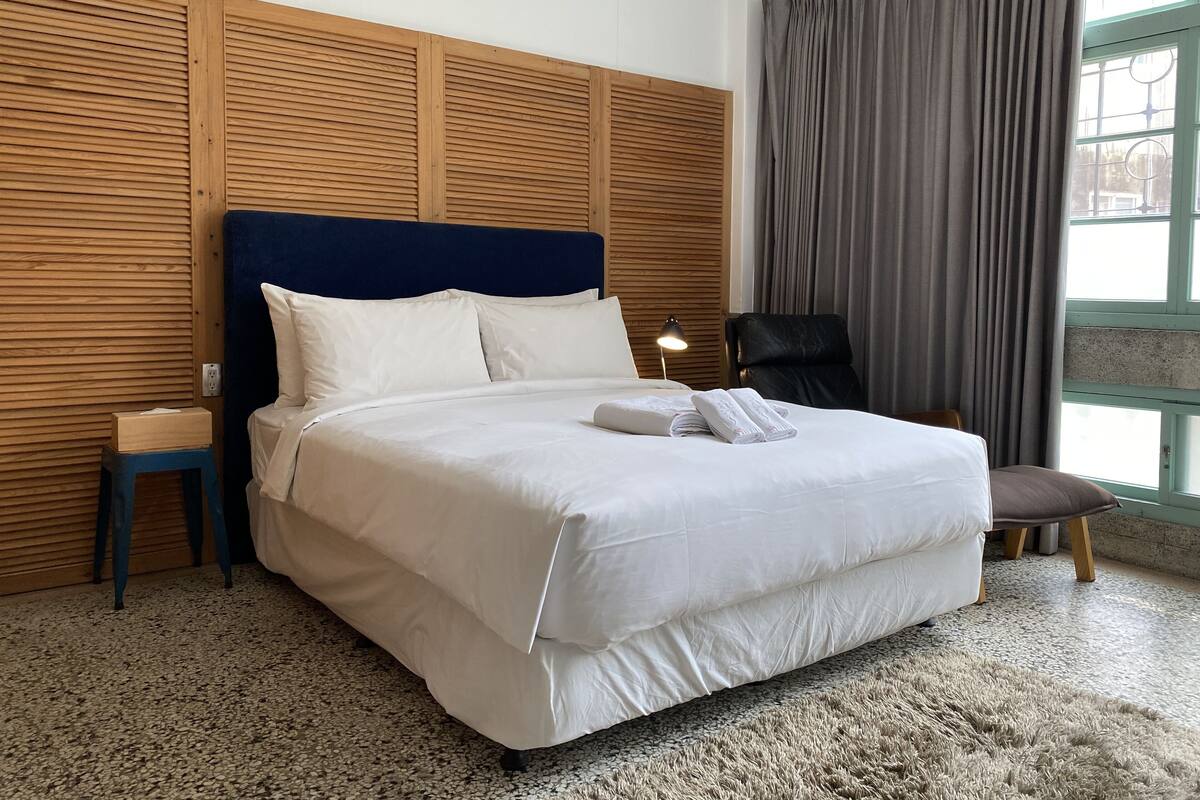 A neatly made bed with white linens, a wooden headboard, a small bedside table, a chair, and a large window with curtains.
