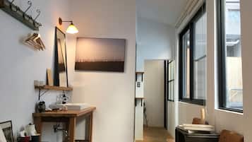 A compact, well-lit room with a wooden desk, a mirror, a shelf with a lamp, and a large window.