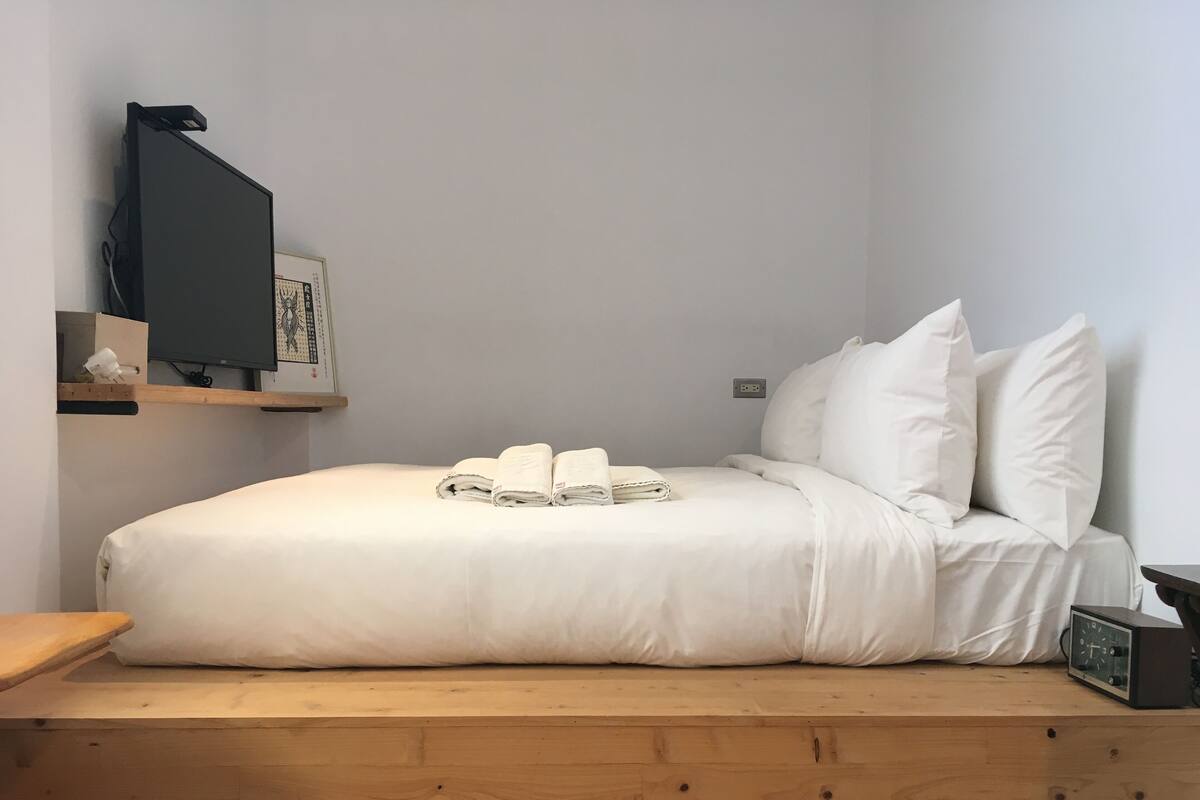 A neatly made bed with white linens and pillows, a bedside table with a clock, and a shelf with a television and other items.