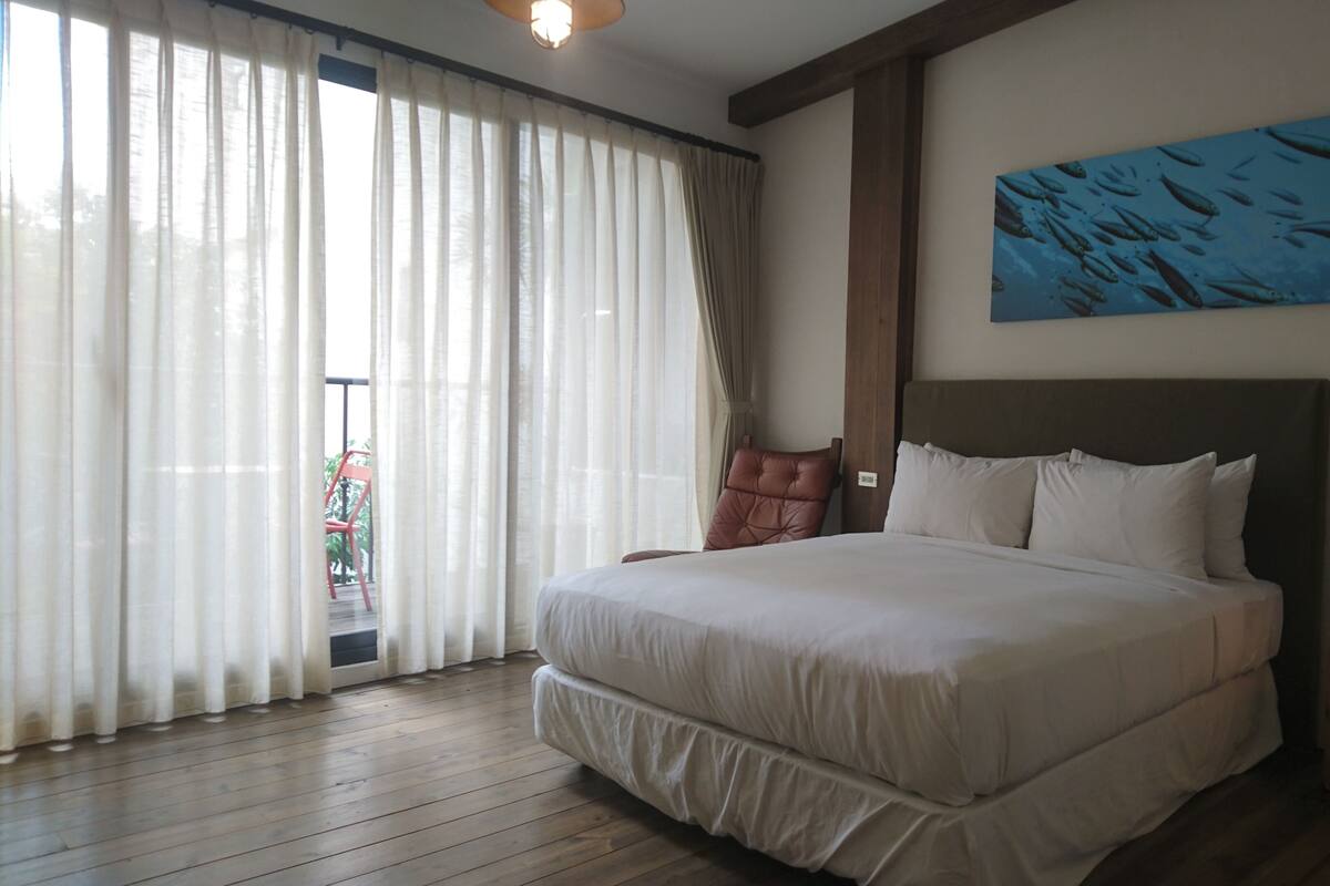 A hotel room with a large bed, a wooden floor, a red chair, a nightstand with a lamp, and a painting on the wall.