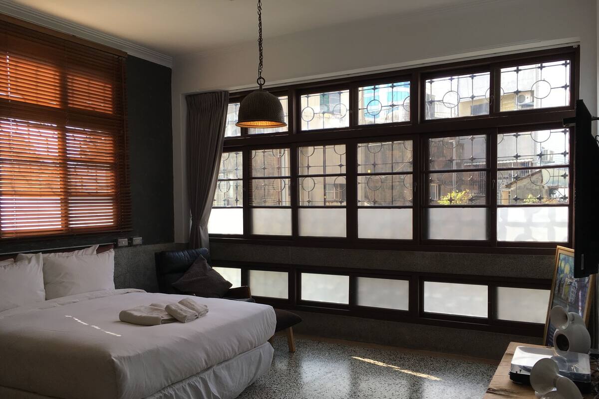 A hotel room with a large bed, a desk, and a view through a window with blinds.