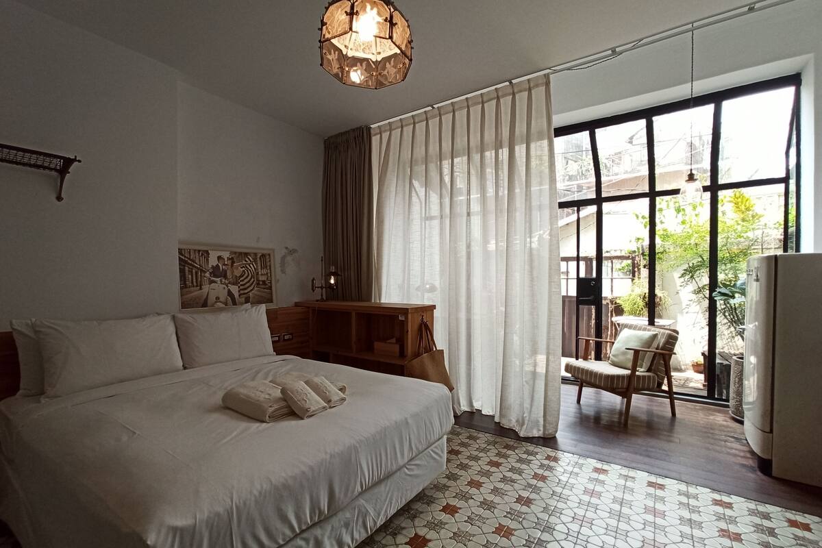A hotel room with a large bed, a desk with a lamp, a chair, and a view of the outdoors through large windows.