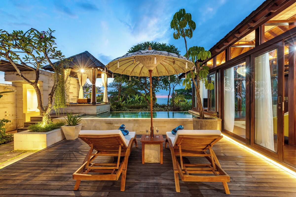 A wooden deck with lounge chairs and a table, overlooking a swimming pool and lush greenery.