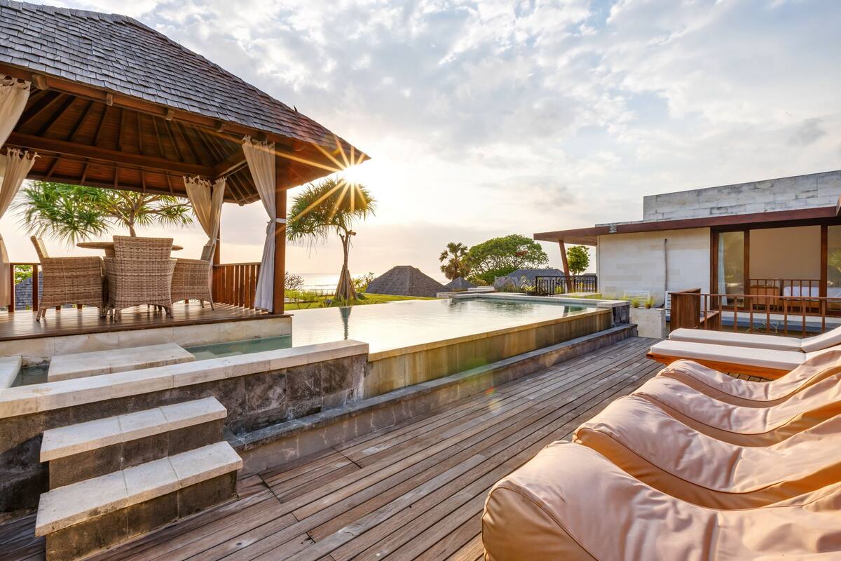 A poolside area with lounge chairs, a wooden deck, and a covered seating area with a view of the pool and palm trees.