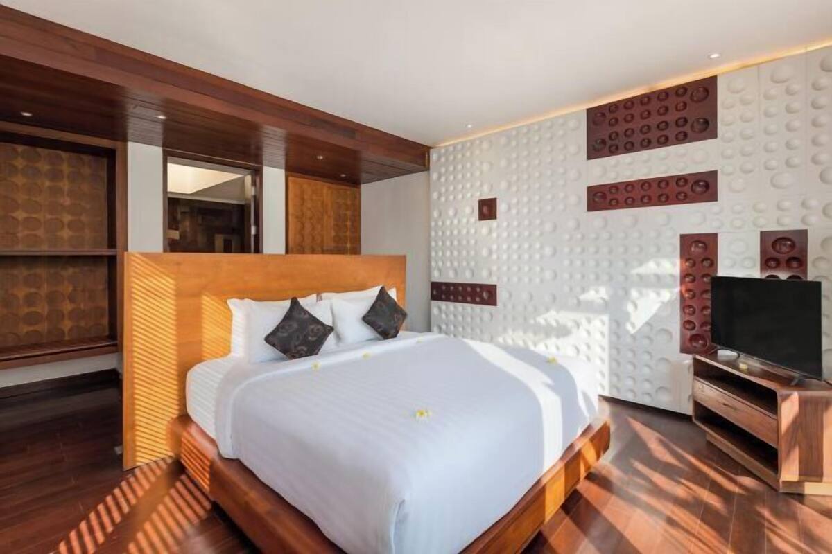 A modern hotel room with a large bed, wooden headboard, a flat-screen TV, and a decorative wall panel.