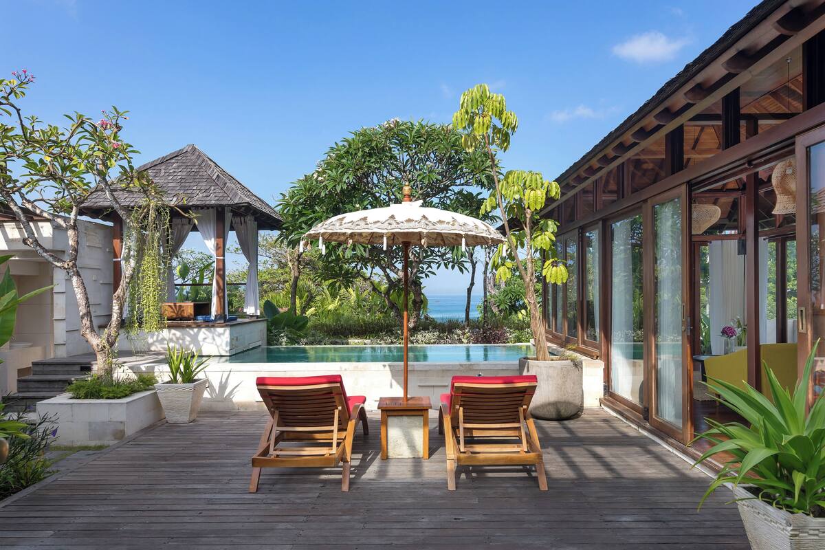 A wooden deck with two wooden lounge chairs and a table, overlooking a pool and ocean view.