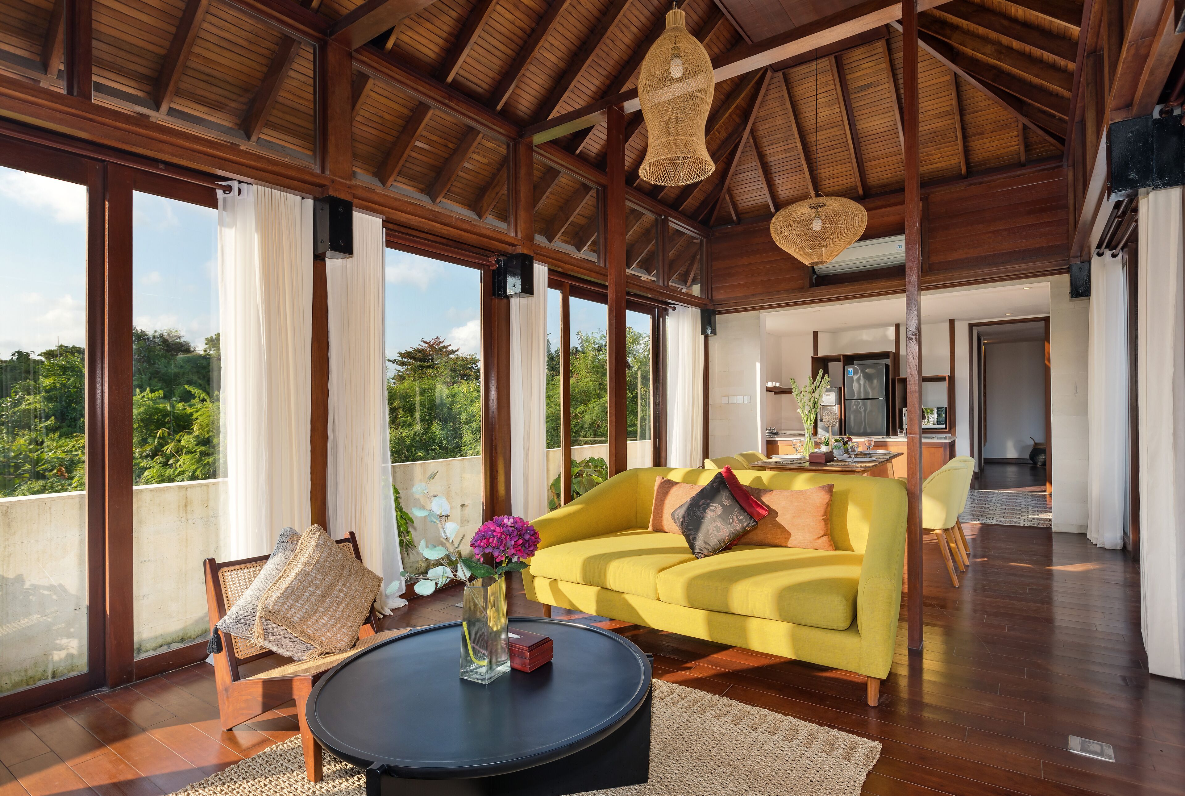 A spacious living area with a yellow sofa, a round coffee table, and a view of lush greenery through large windows.