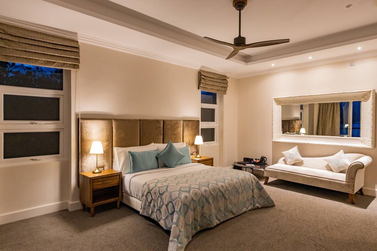 A bedroom with a bed, bedside tables, a nightstand, a sofa, a ceiling fan, and a large mirror.