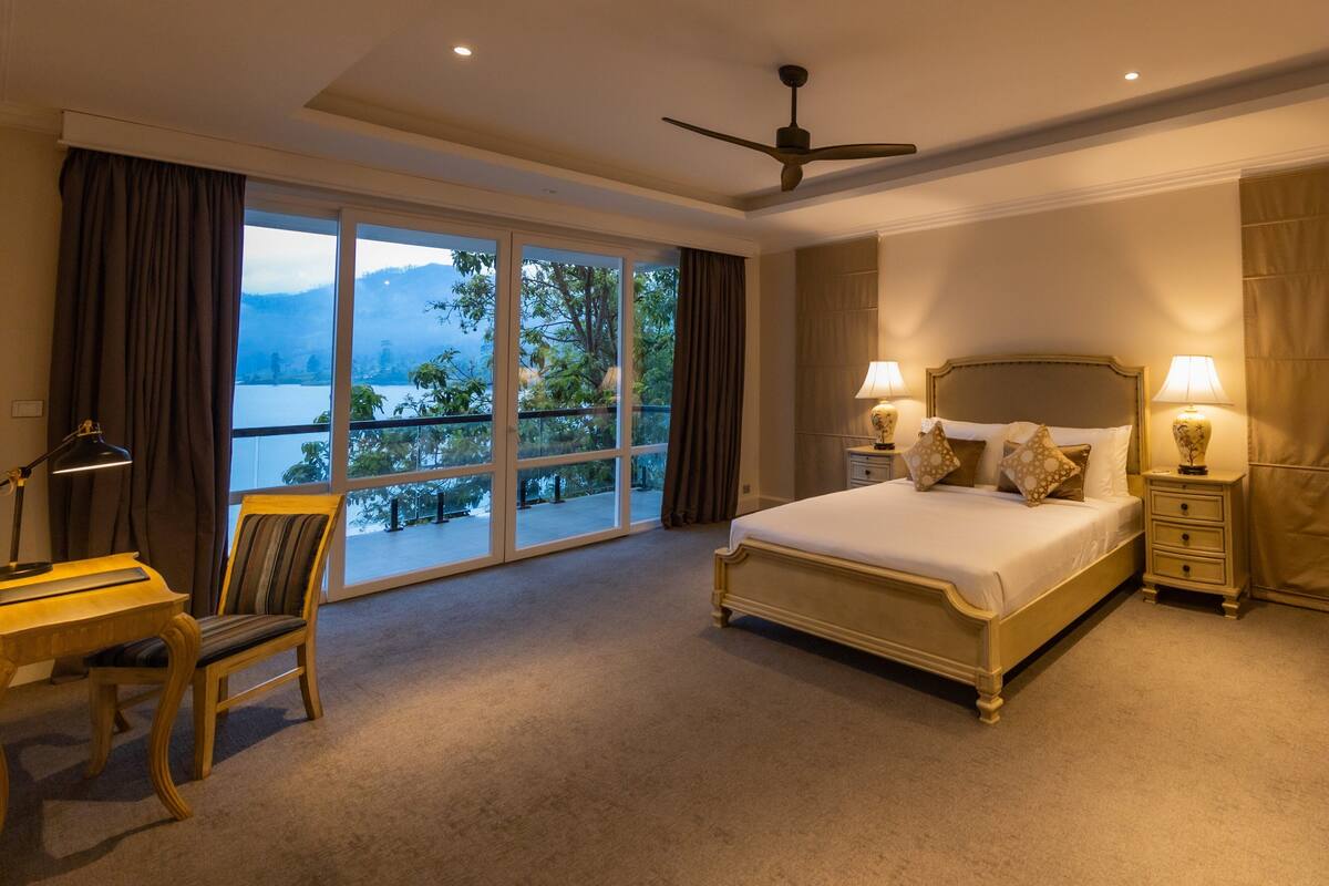 A bedroom with a large bed, a desk, a chair, and a view of a lake and mountains.