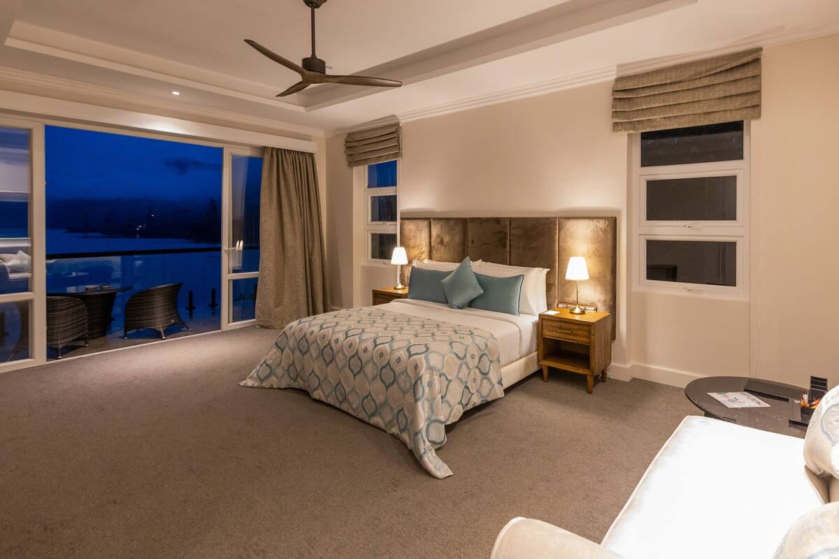 A bedroom with a bed, two bedside tables, a nightstand, a ceiling fan, a large window with curtains, and a view of the outside.