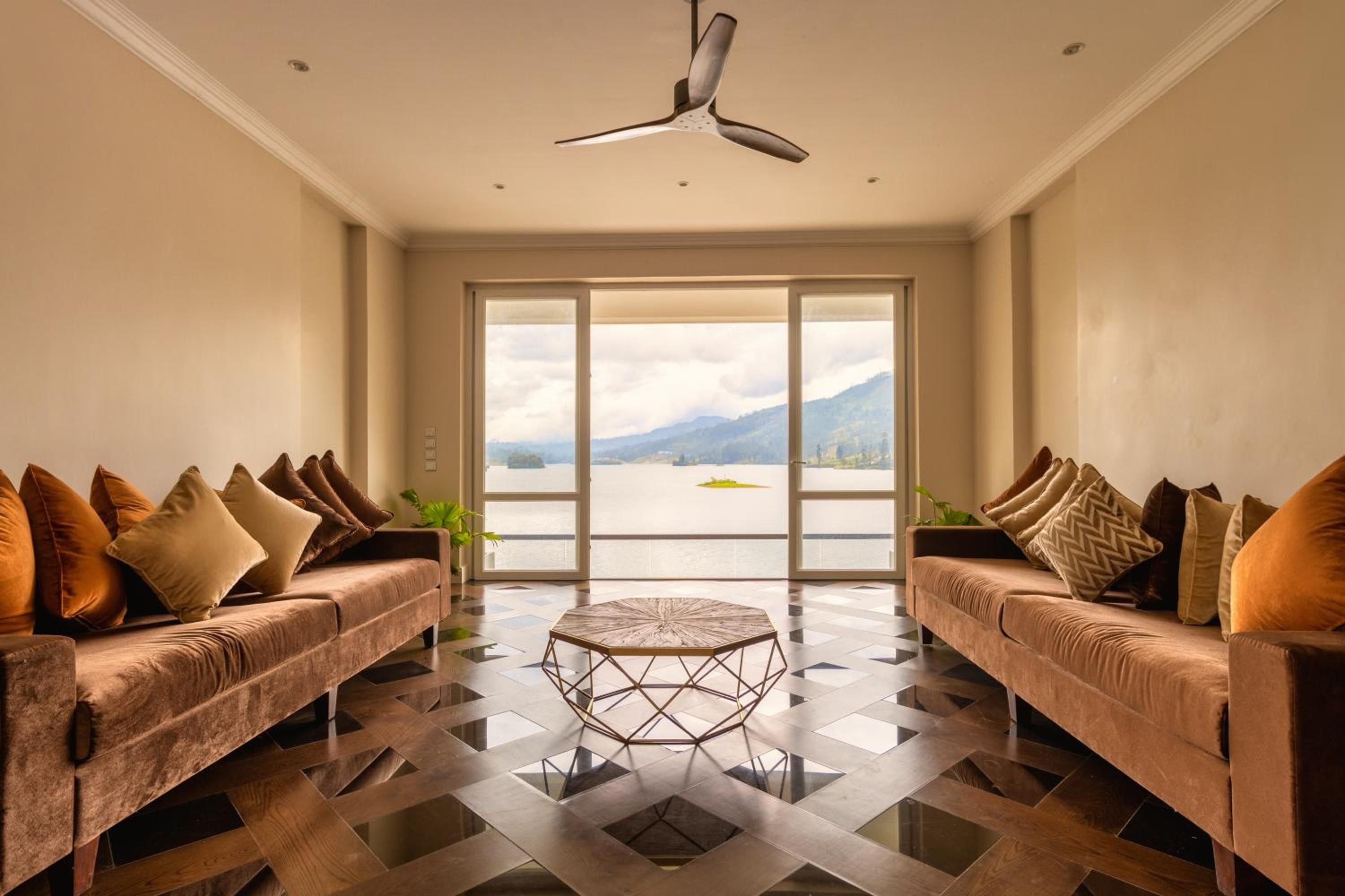 Una sala de estar moderna con dos sofás, una mesa de centro y vistas a un lago y montañas a través de amplios ventanales.