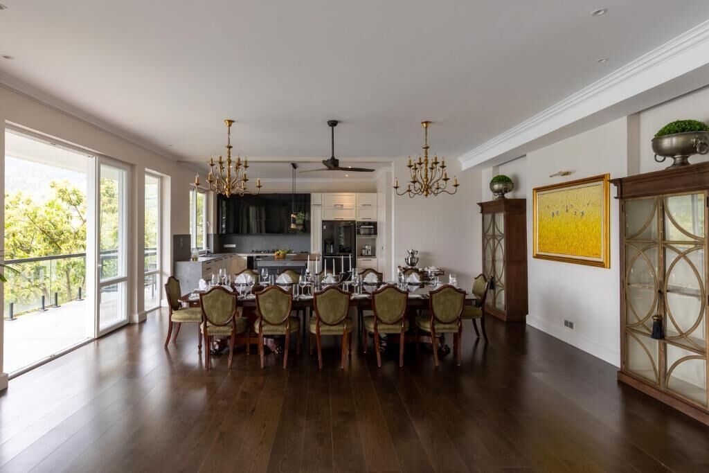 Un comedor amplio con una larga mesa dispuesta para seis personas, rodeada de sillas de madera. Se ve una cocina moderna al fondo, y un ventanal grande proporciona luz natural.