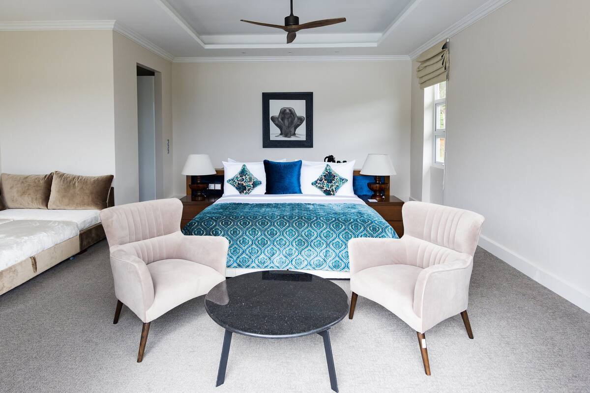 A hotel room with a bed, two armchairs, a small round table, a ceiling fan, and a framed picture on the wall.