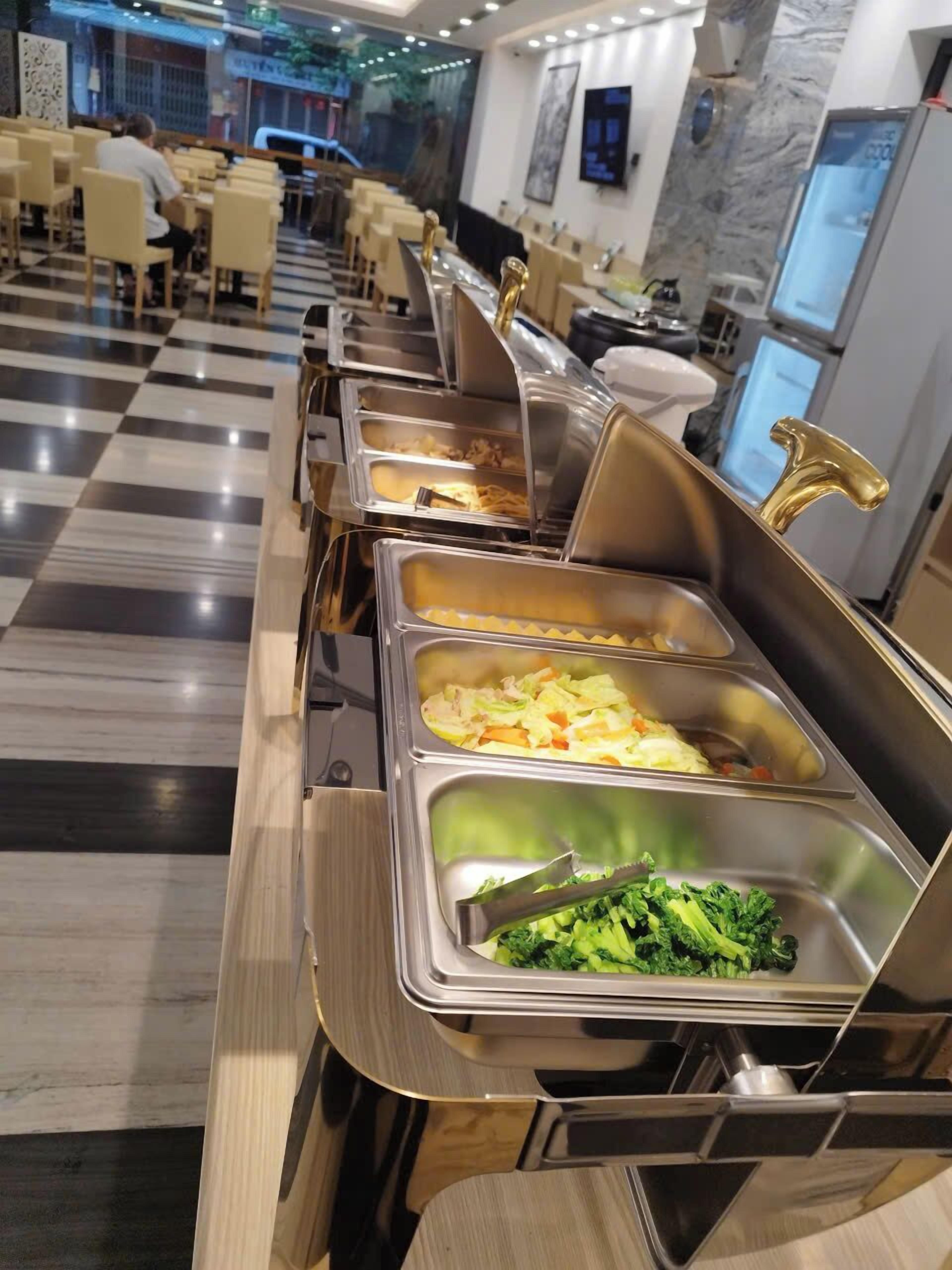 Ein Buffet mit verschiedenen Speisen in Edelstahl-Warmhaltebehältern, darunter Krautsalat und Brokkoli, in einem Essbereich mit Schachbrettboden und Marmorwänden.