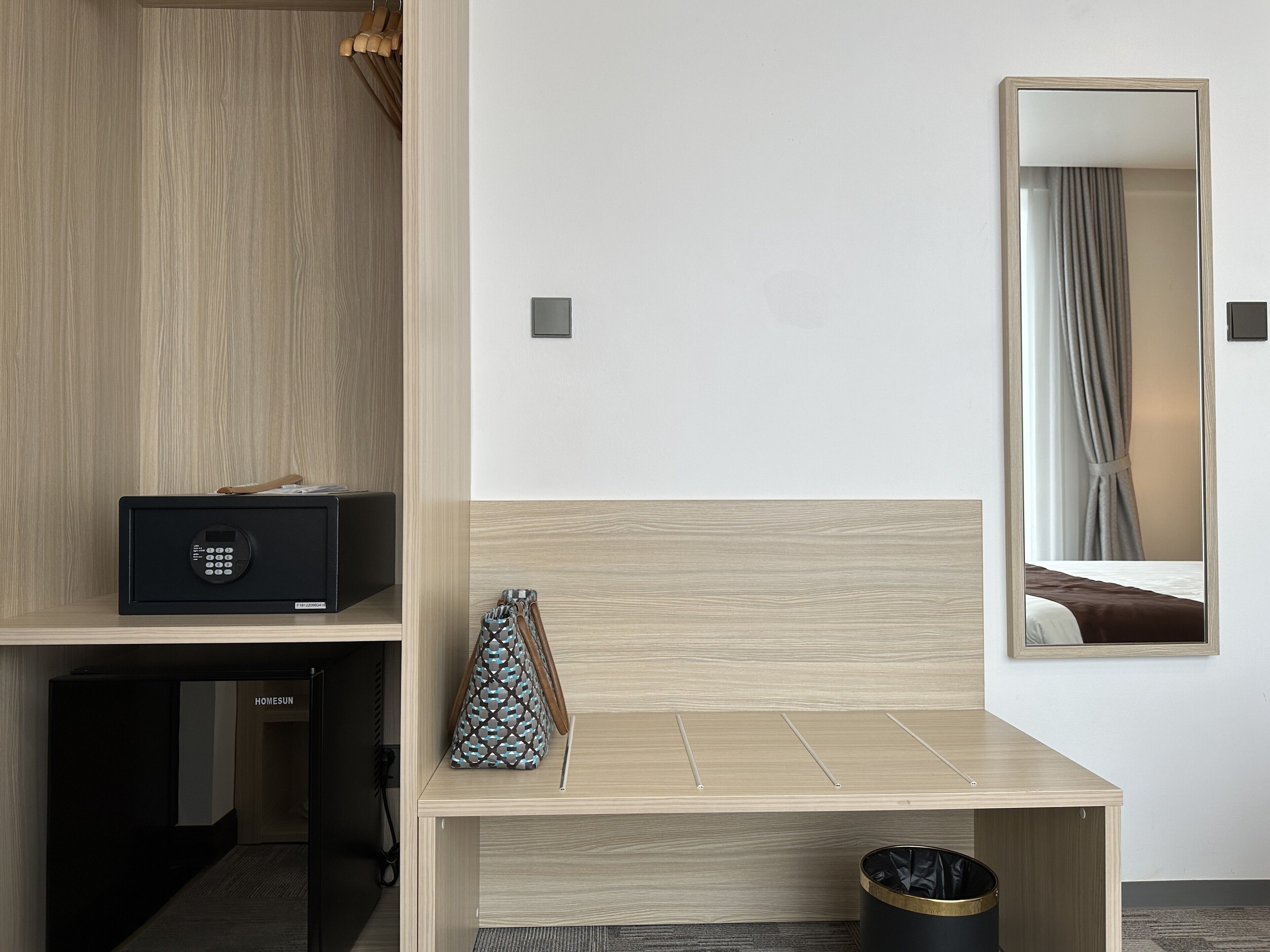 A hotel room with a wooden bench, a small table, a trash can, a wall-mounted mirror, and a door leading to another room.