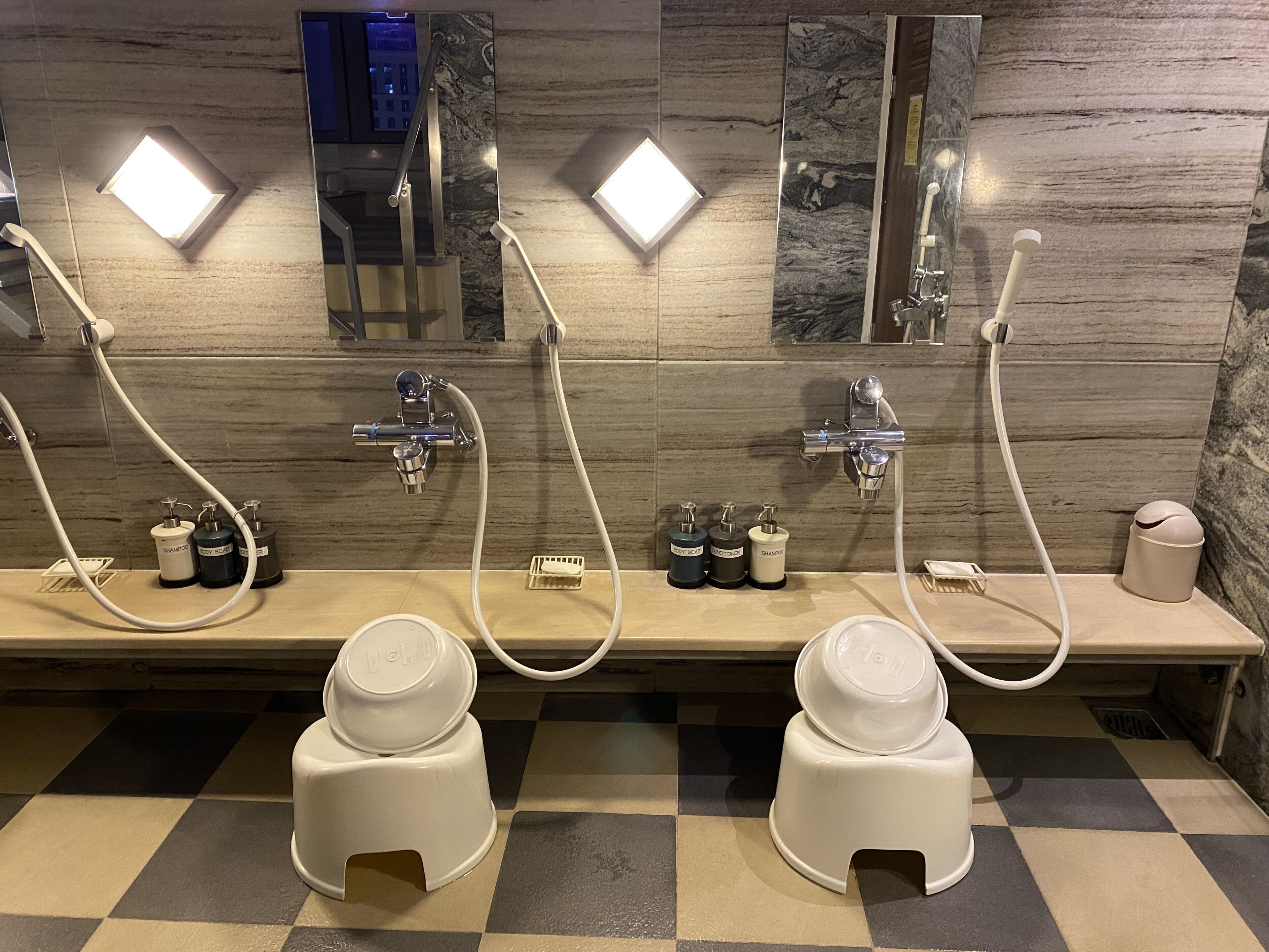 Baño público con dos lavamanos, cada uno con cabezal de ducha manual, y un espejo arriba.
