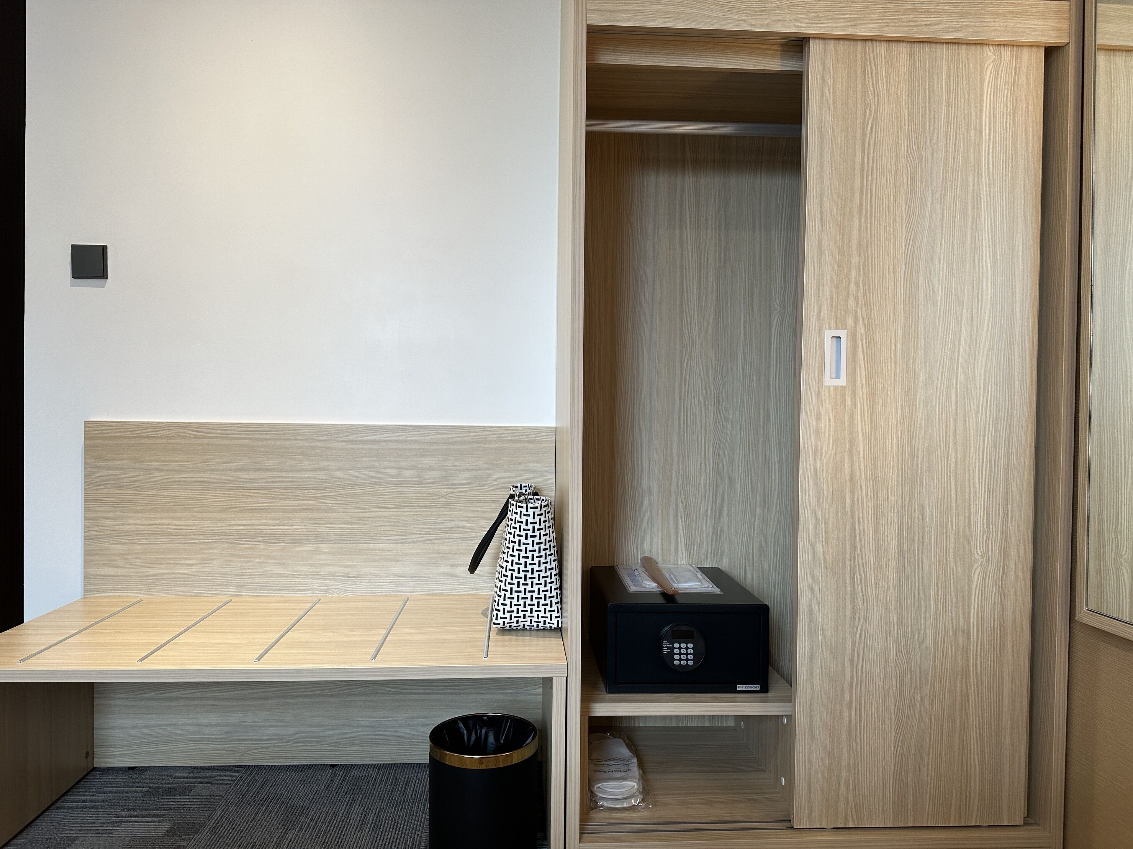 Ein Hotelzimmer mit einer hölzernen Bank, einer schwarzen Mülltonne, einem schwarzen Safe und einer gemusterten Tasche auf der Bank.