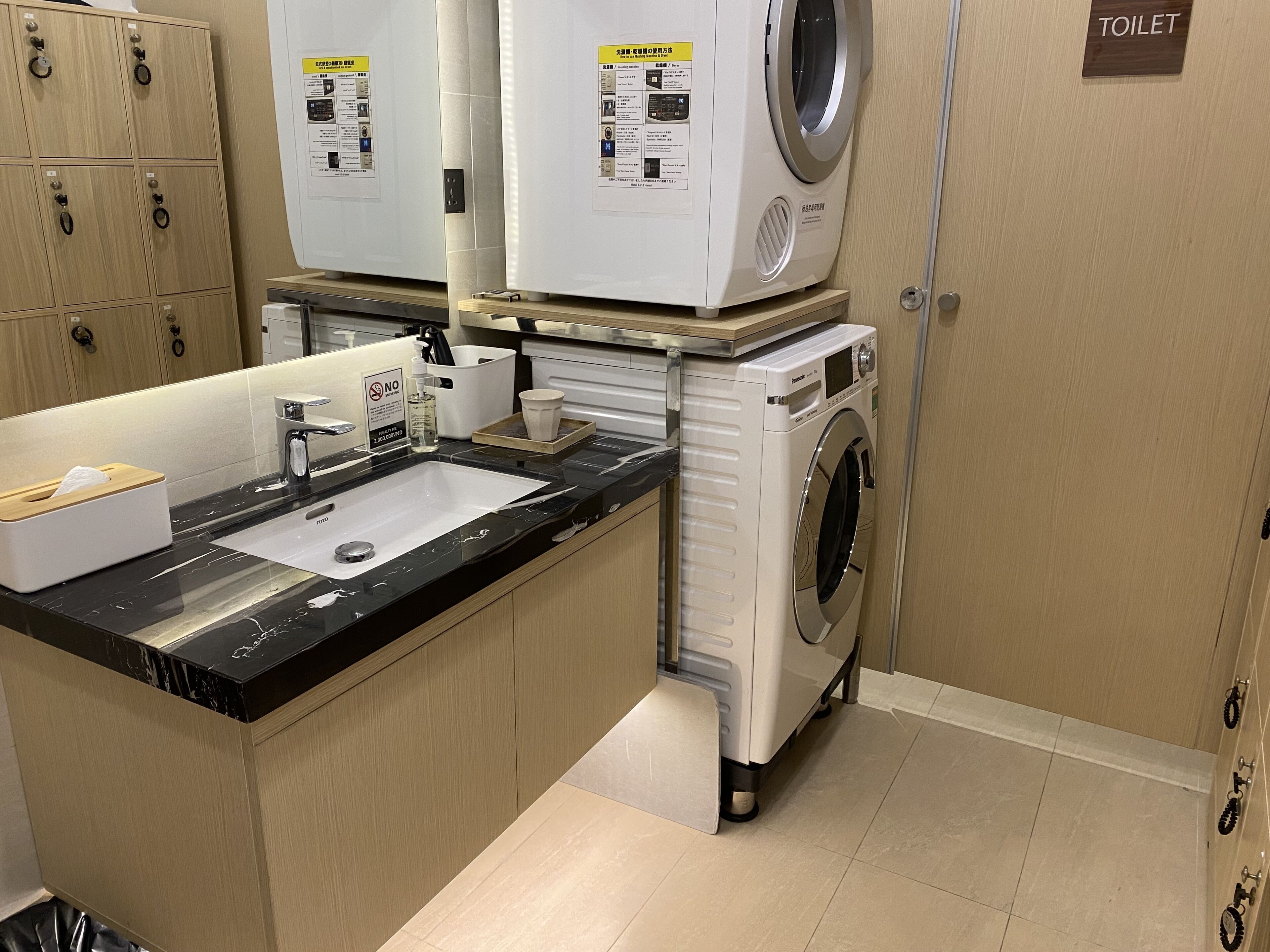 A hotel bathroom with a sink, two washing machines, a toilet, and wooden cabinets.