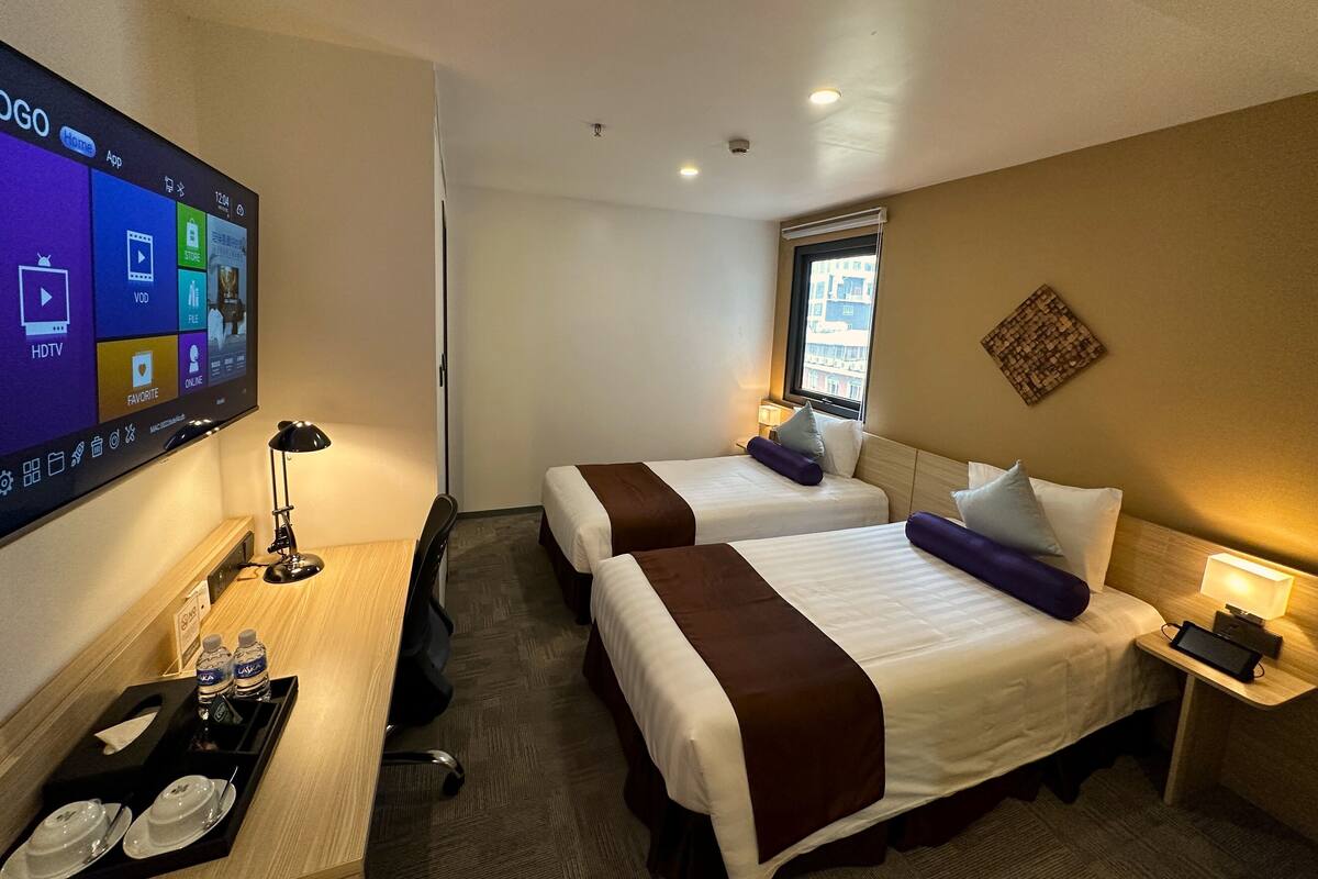 Une chambre d’hôtel avec deux lits, un bureau avec un ordinateur, un téléviseur affichant un écran d’accueil et une fenêtre donnant sur des immeubles.