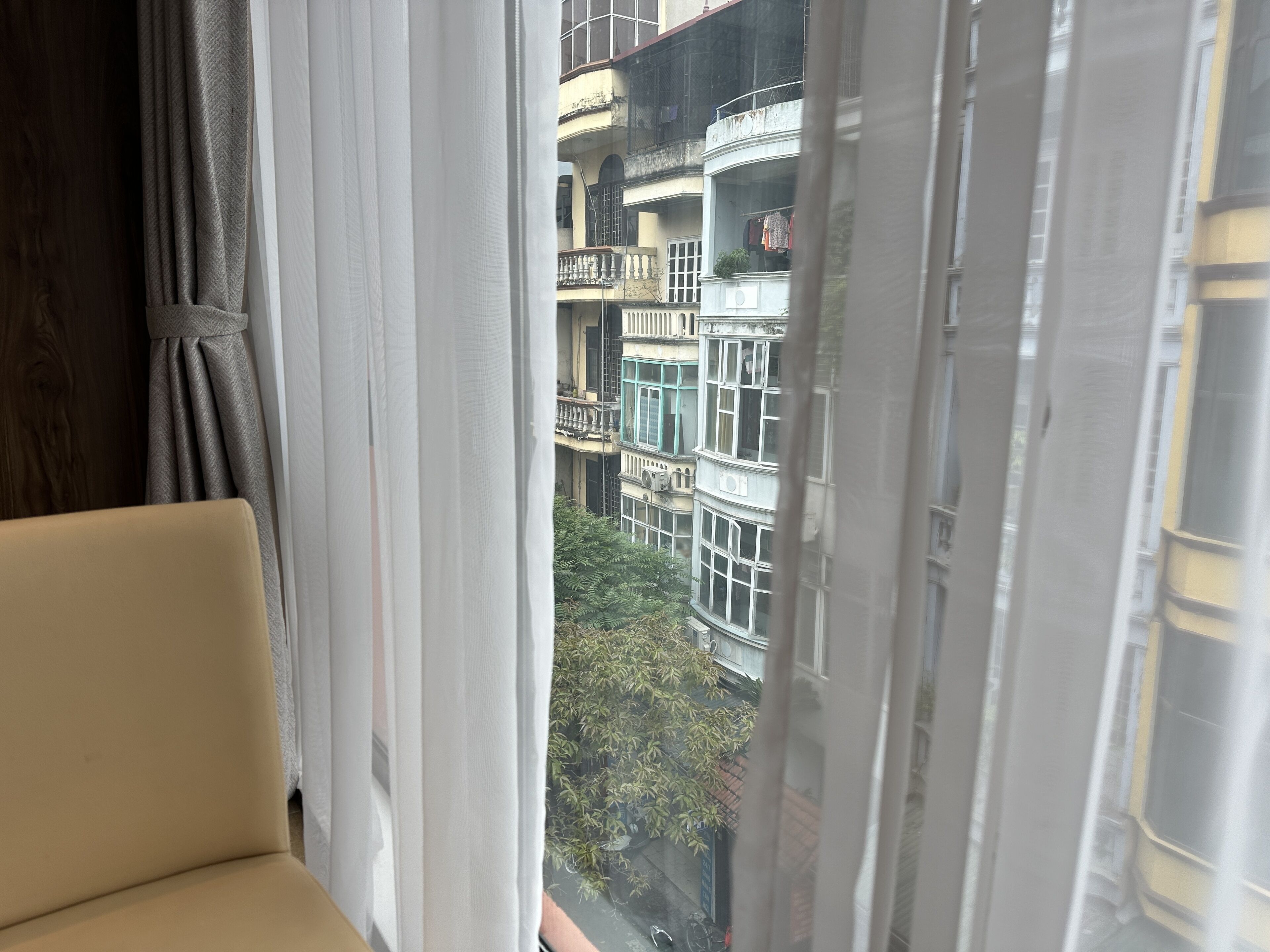 Une vue par la fenêtre, à travers les rideaux, dévoilant un immeuble à plusieurs étages, doté de balcons et surplombant un espace verdoyant.