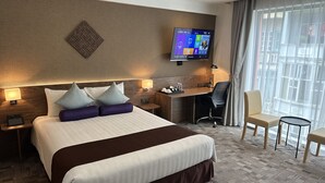 Habitación de hotel con una cama grande, un escritorio, una silla, un televisor montado en la pared y una ventana con cortinas.