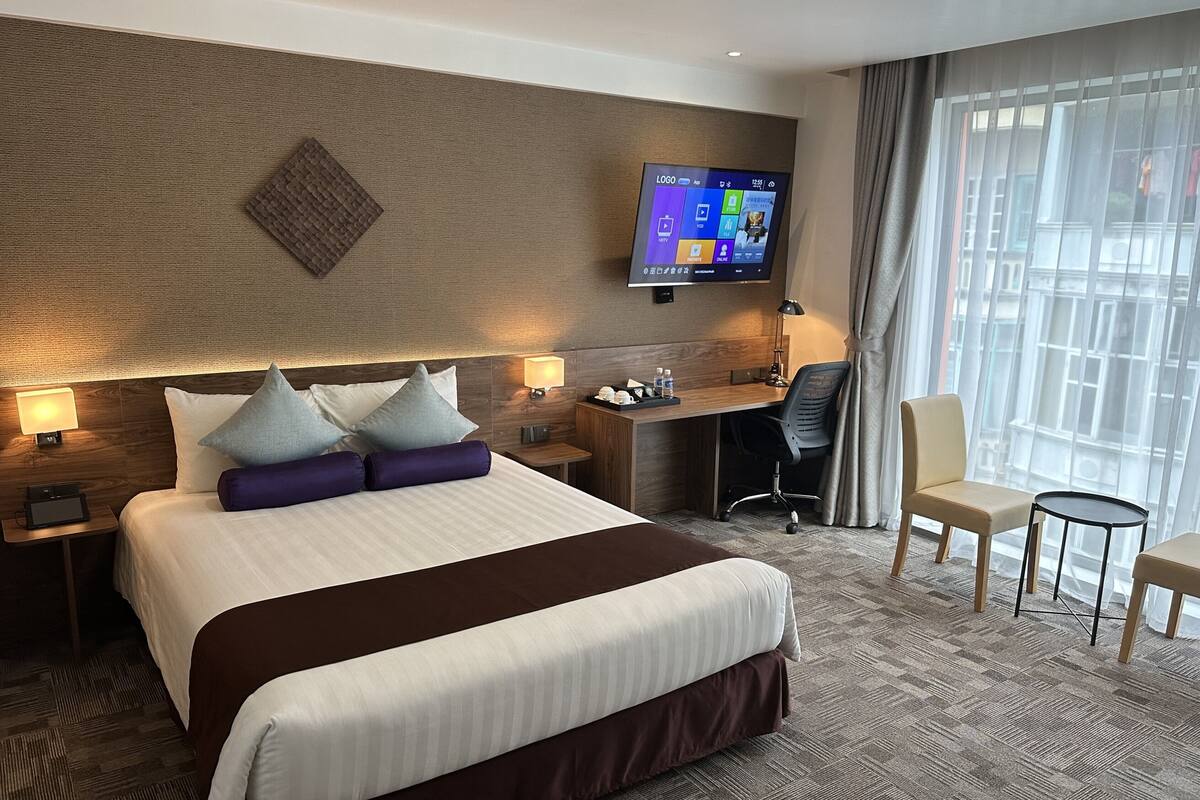 Une chambre d’hôtel avec un grand lit, un bureau, une chaise, un téléviseur fixé au mur et une fenêtre avec des rideaux.