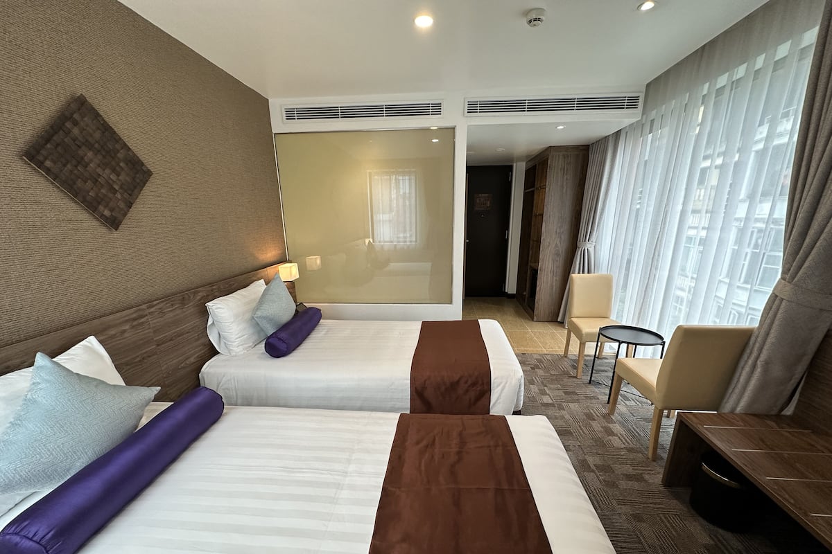 Une chambre d’hôtel avec deux lits, un coin salon comprenant une chaise et une petite table, et de grandes fenêtres avec des rideaux.
